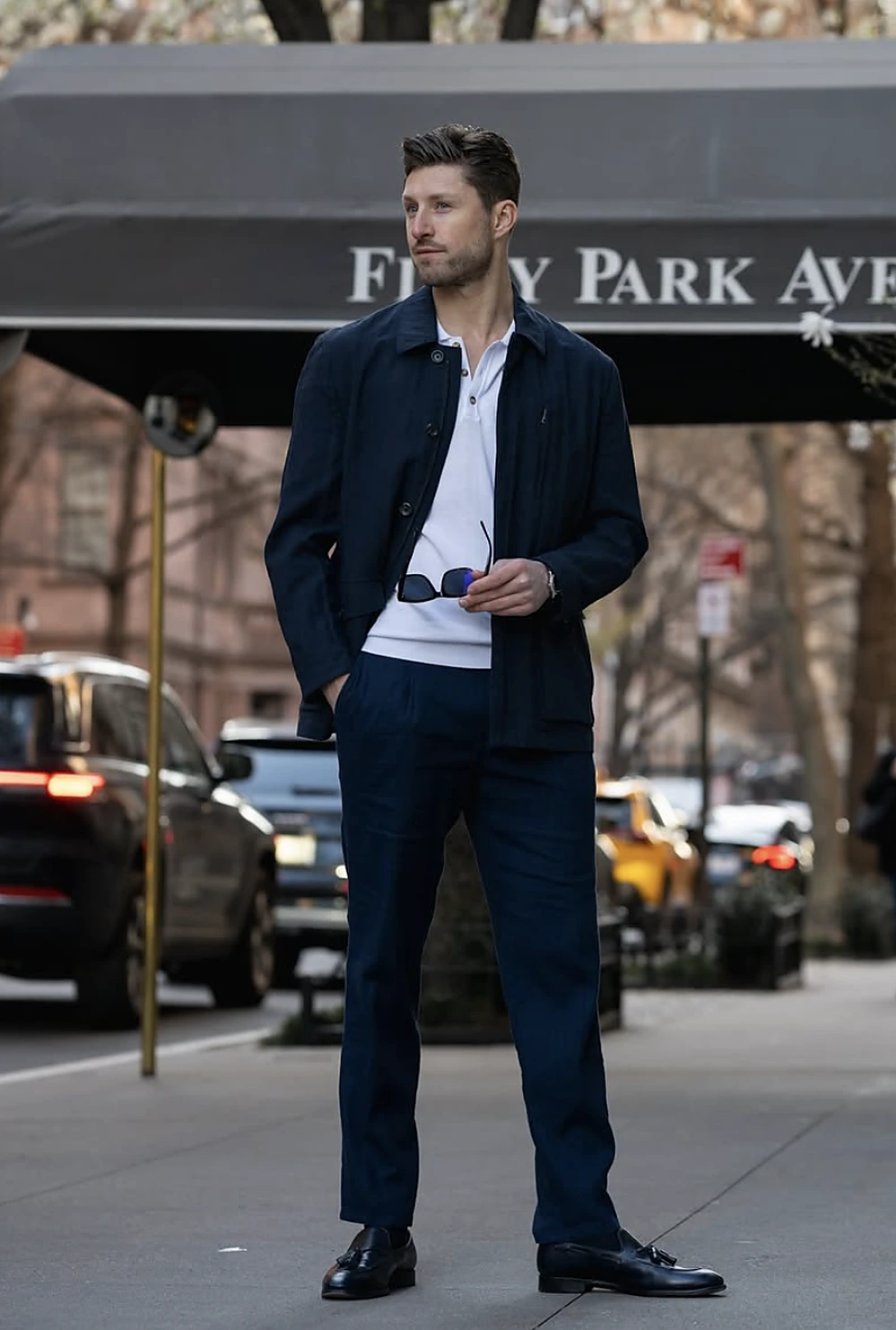 Tall man wears a navy overshirt with matching slim pants, white polo, black shoes, and sunglasses outdoors.