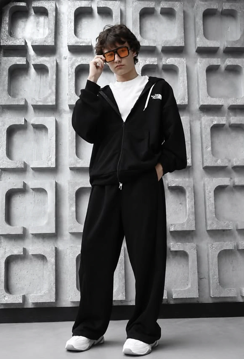 Young man in black zip hoodie, baggy black pants, white tee, orange-tinted glasses, and white sneakers.