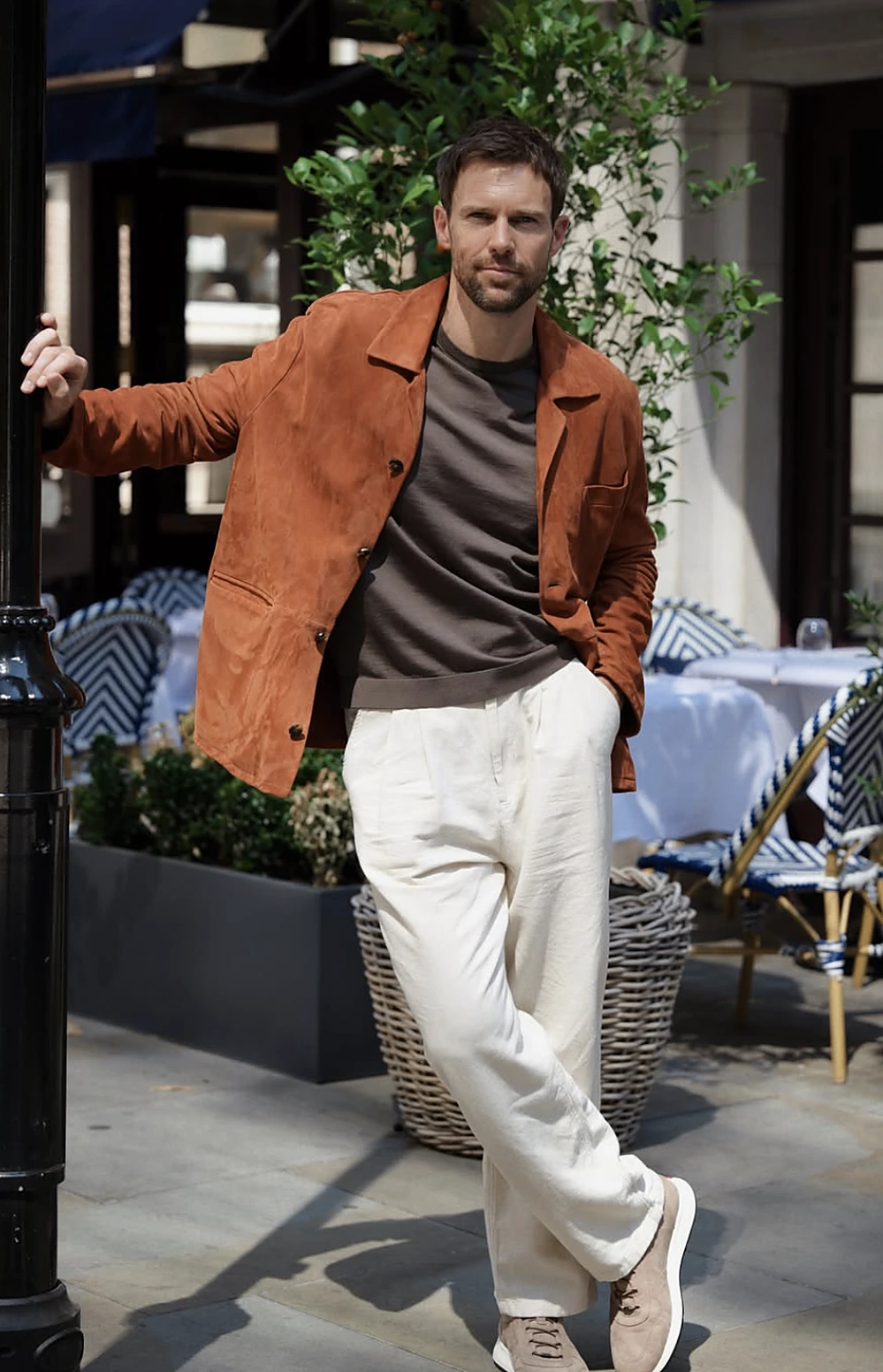 Man wearing a rust suede jacket, dark knit top, cream trousers, and tan sneakers beside outdoor café seating.